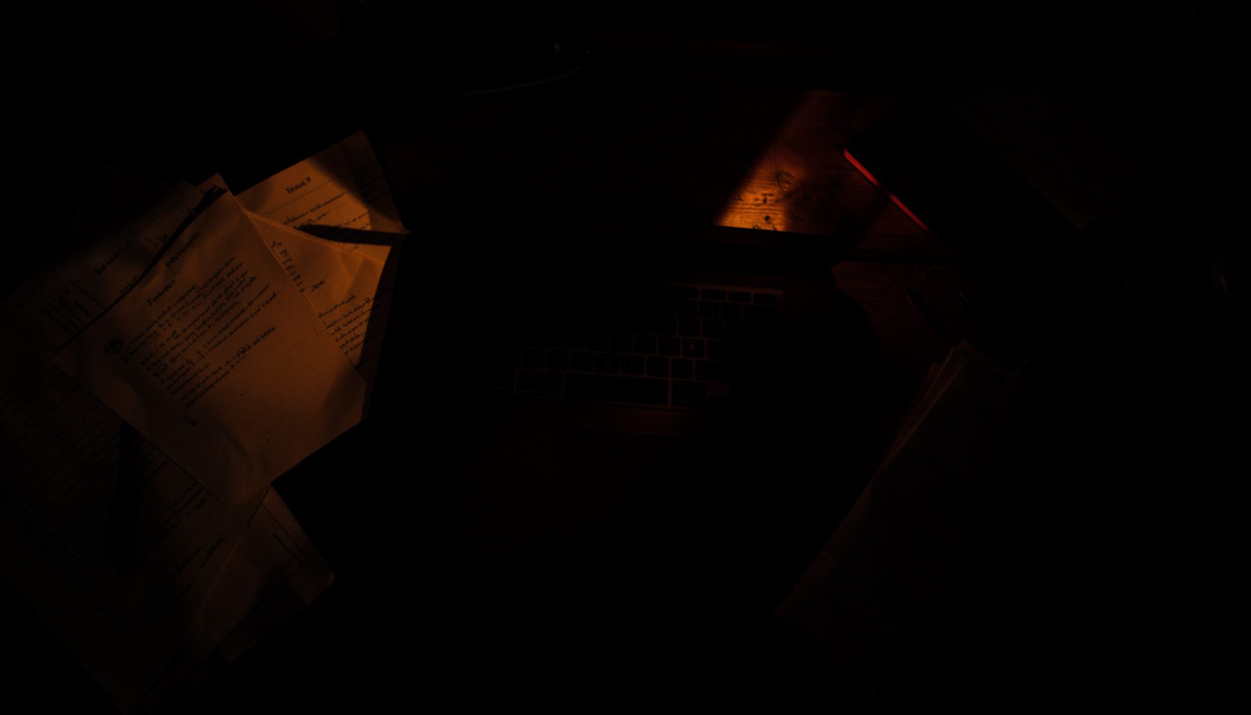 A dark, low-key overhead photograph of a laptop on a wooden desk bathed in deep shadow, with warm amber light spilling across scattered papers, evoking a quiet late-night SEO planning session.