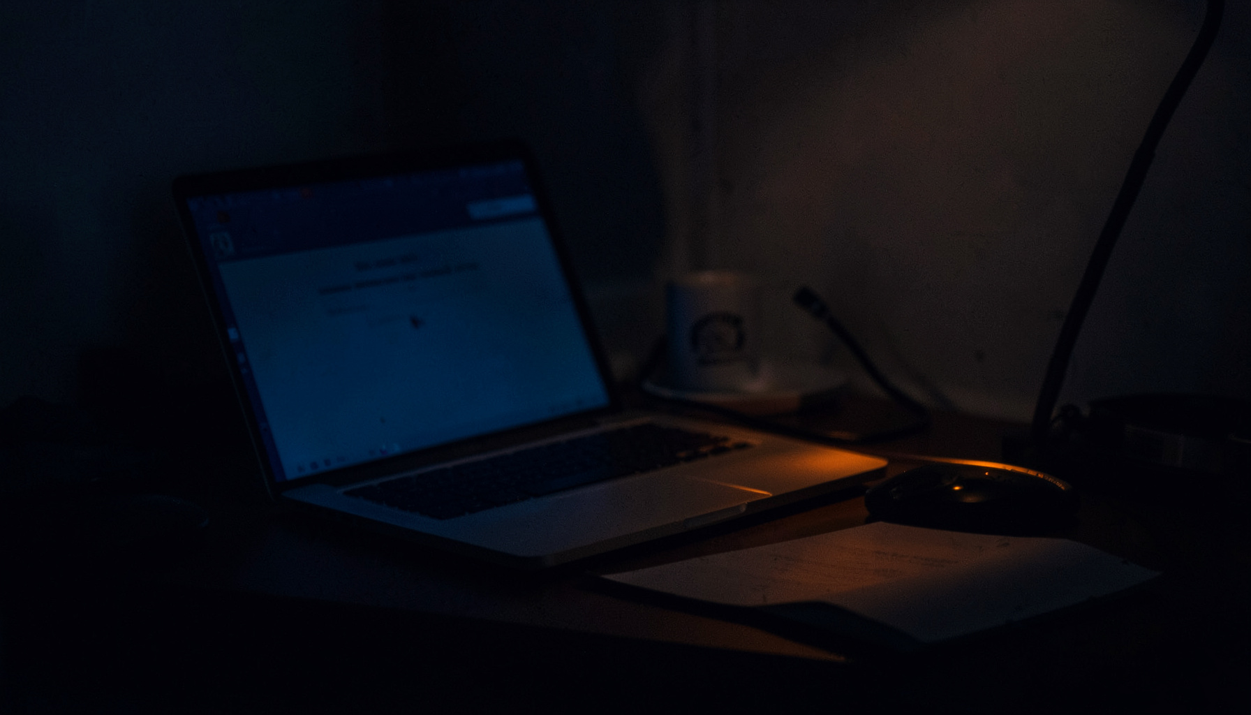 A dark, moody desk scene with a laptop screen glowing faintly in low light, deep shadows across the table, muted navy and amber tones suggesting focused late-night work