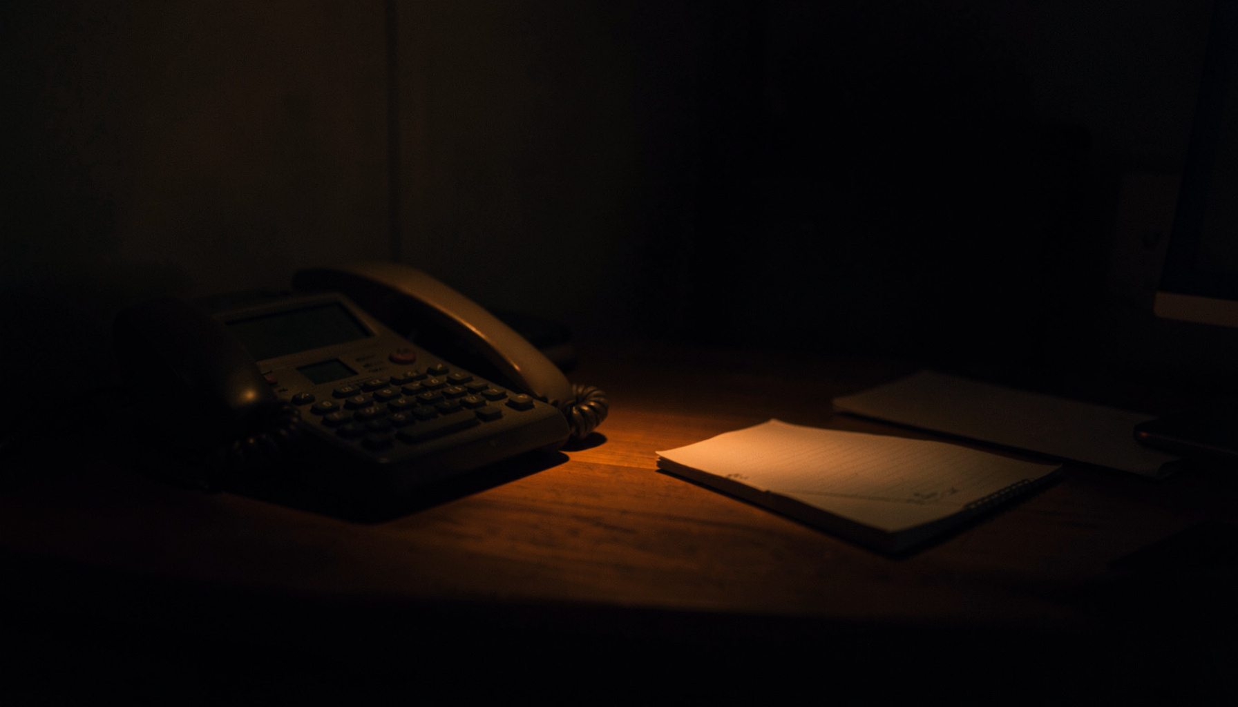 A dim, low-key photograph of a softly lit office desk with a telephone and notepad in deep amber and slate tones, evoking a calm professional mood