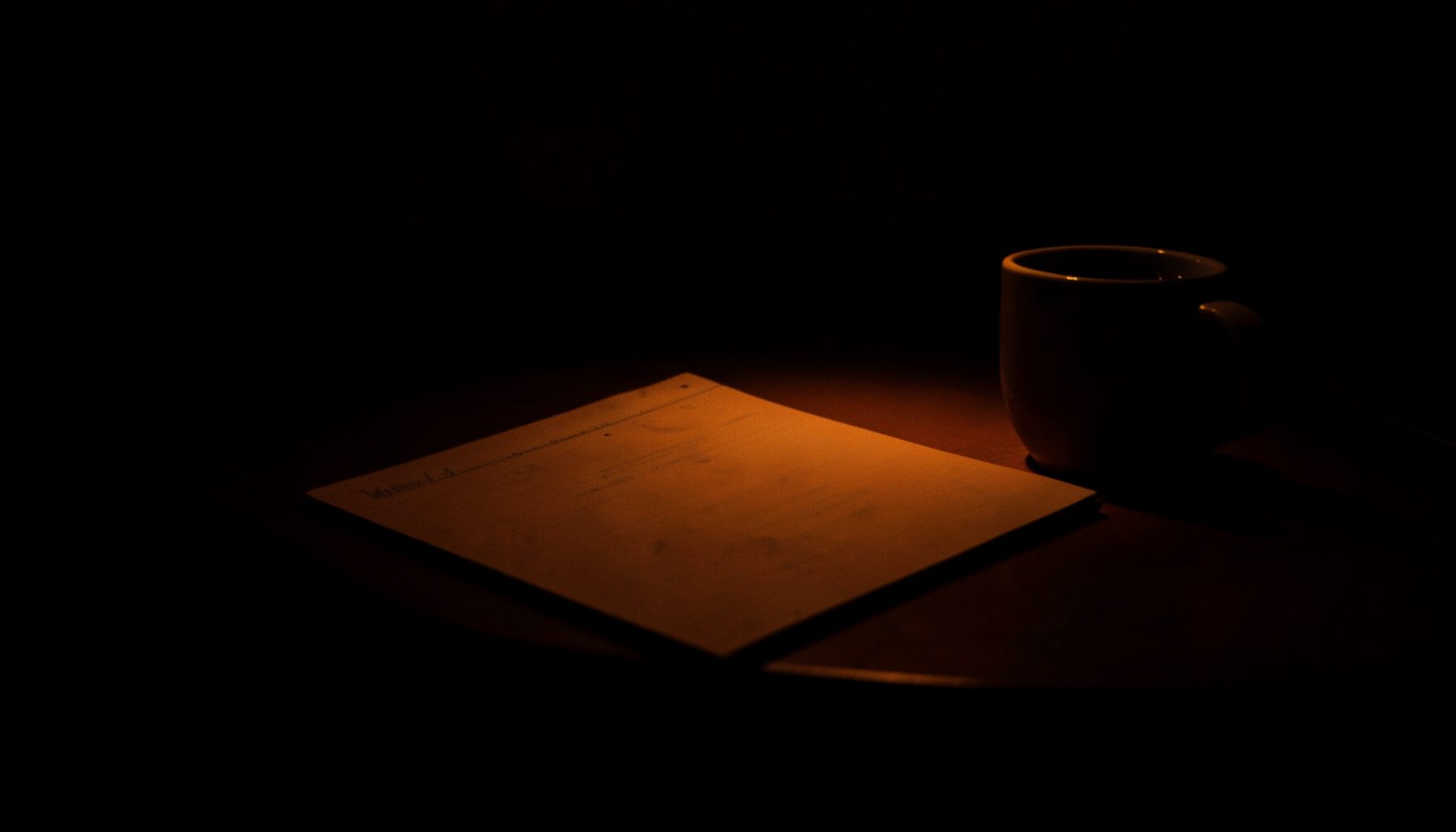 Dark low-key photograph of a desk at night with deep shadows and warm amber highlights on a notebook and coffee cup